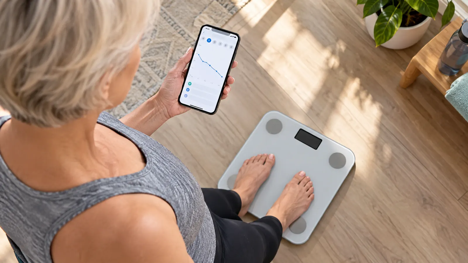 A hand holding a smartphone showing a graph of weight trends next to a smart scale on a bathroom floor